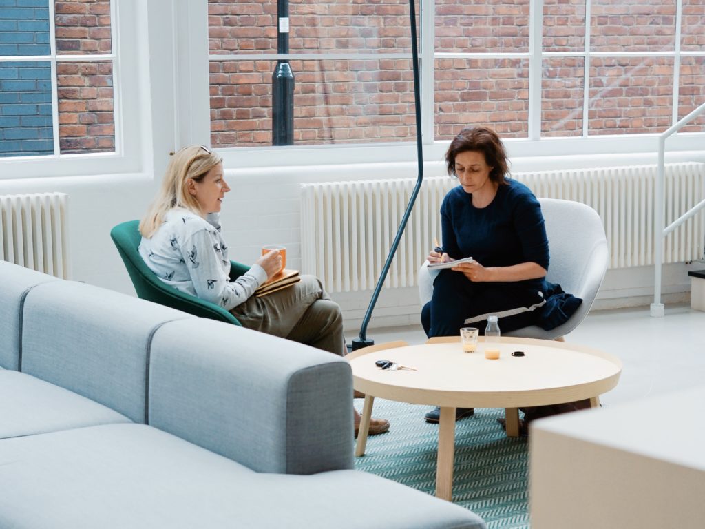 Two women are sitting down in a bright room, taking notes and collaborating.