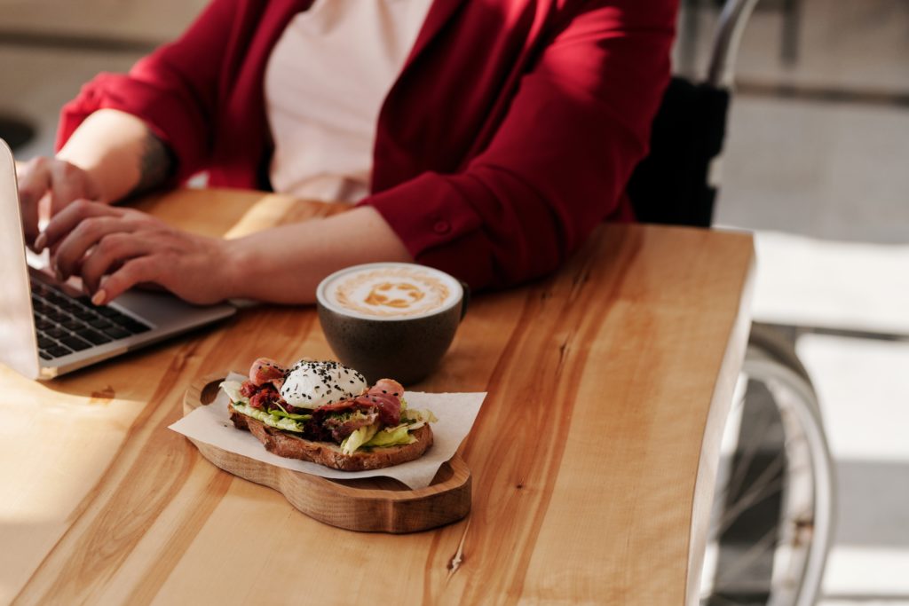 A woman is sitting down at a table with her laptop. A avocado toast and a cup of latte coffee are next to her computer.