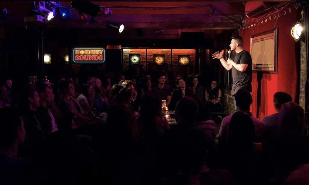 A standup comedian is doing a show in a dimmed-light room in front of an audience.