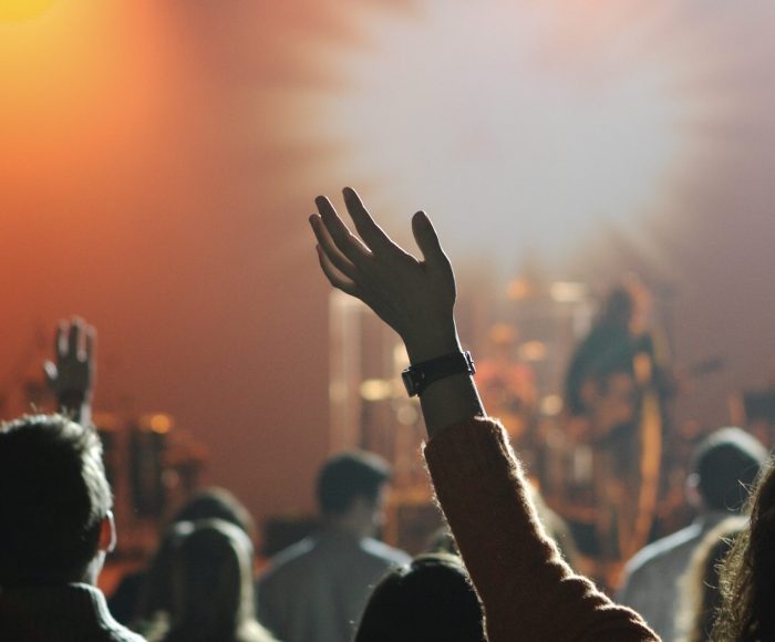 A band is playing on stage in the background as the camera focuses on hands up in the air from the audience.